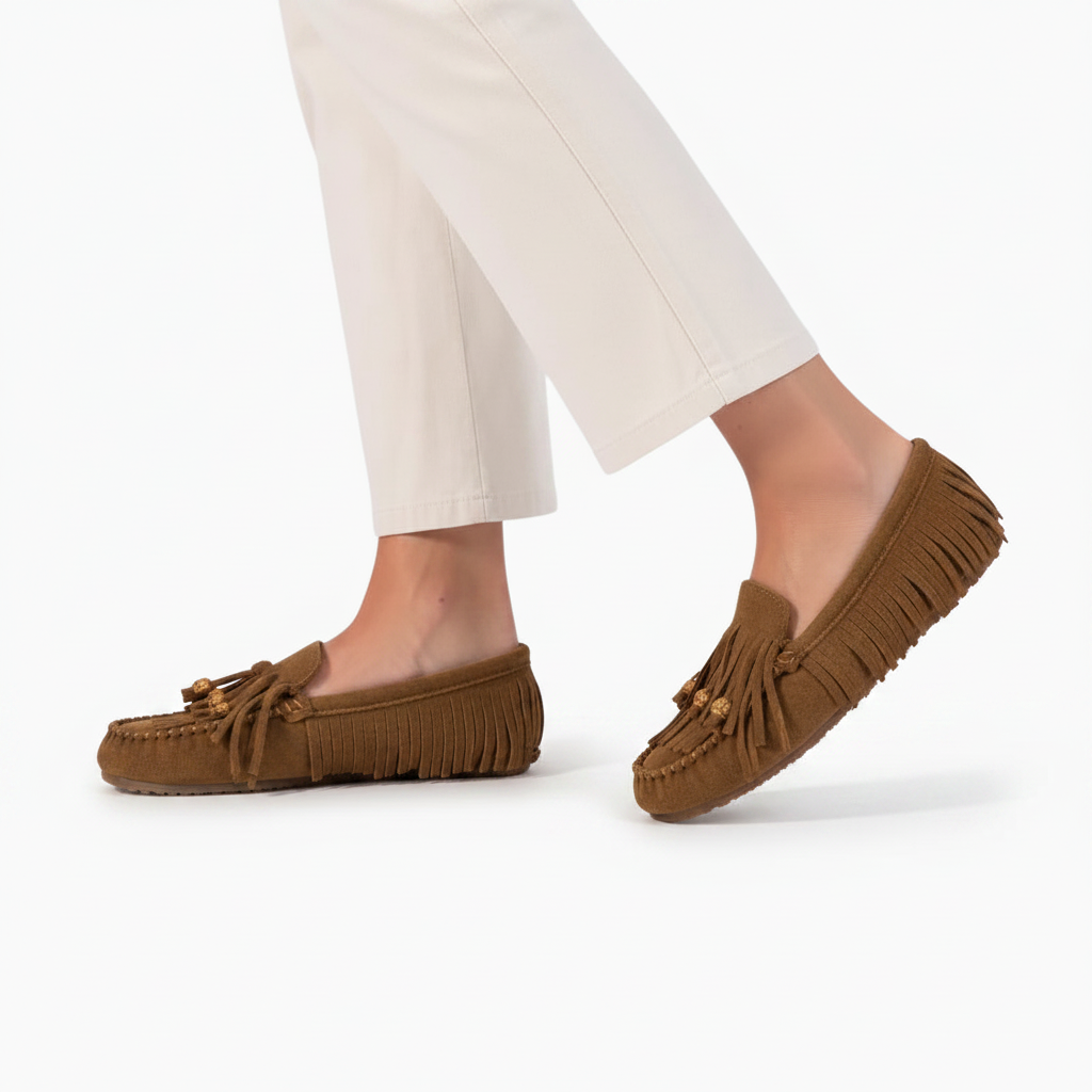 Pied féminin chaussé d'un mocassin en daim marron à franges, posé élégamment sur le sol.