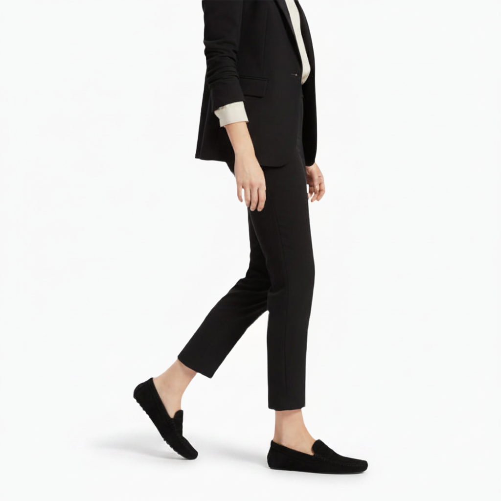 Femme assise, croisant les jambes, ses mocassins en daim noir complétant une jupe fluide avec subtilité.