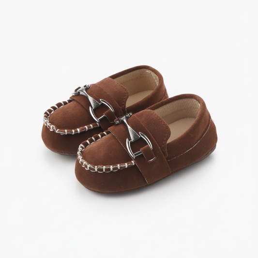 Mocassin bébé en daim marron avec chaînette dorée, posé sur un tapis doux et moelleux.