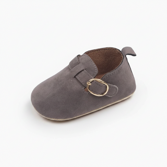 Mocassin bébé gris en daim, idéal pour les premiers pas, délicatement posé sur un plancher en bois clair.