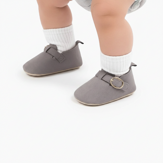 Pieds de bébé confortablement chaussés des mocassins gris en daim, vus de face sur un tapis douillet.