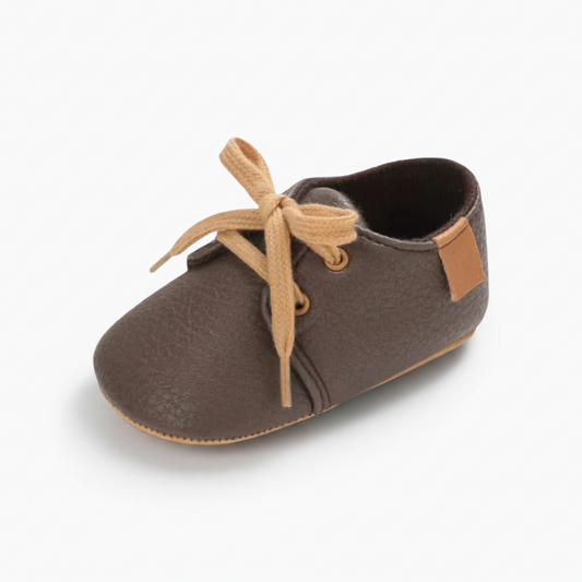 Mocassins bébé en cuir marron foncé, lacets ajustables, posés pour montrer l'élégance classique.